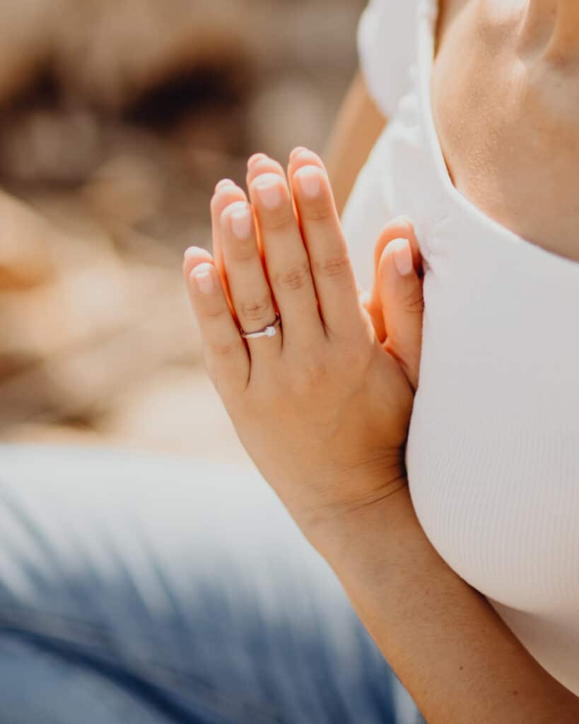 Mains féminines jointes, bague de fiançailles en diamant sur l'annulaire. Peau bronzée, haut blanc, fond flou.