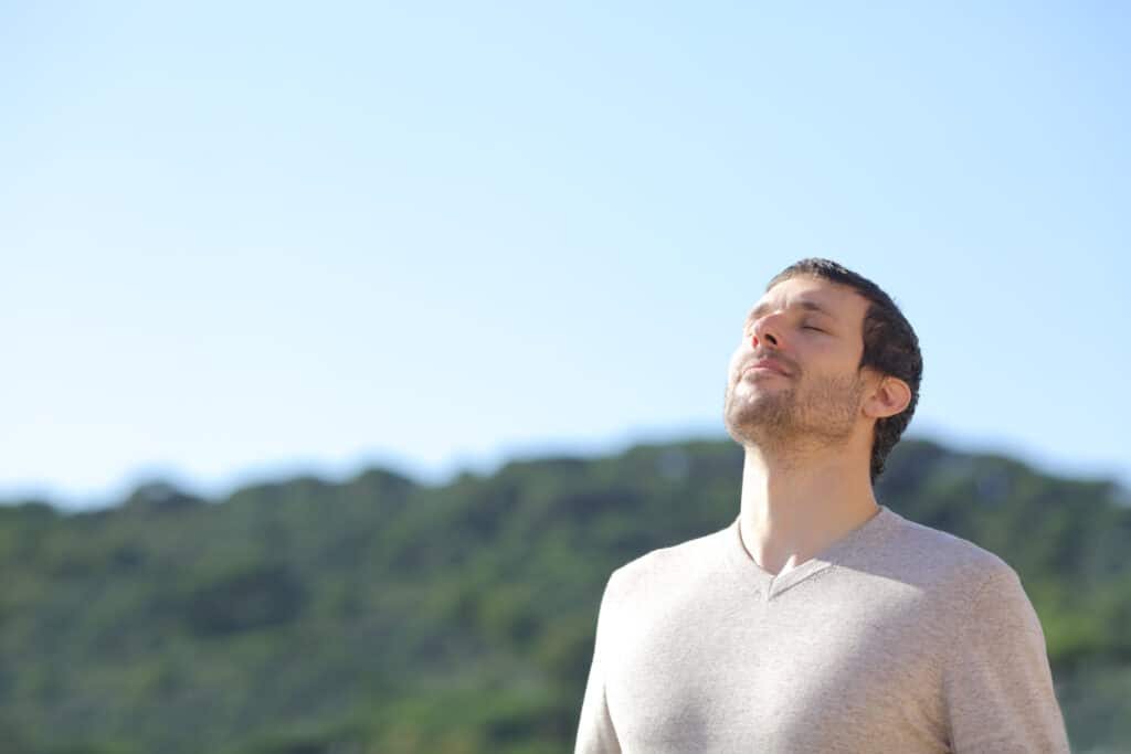 Homme barbu aux yeux fermés, visage levé vers le ciel bleu clair. Il semble se détendre et respirer l'air frais en pleine nature verdoyante.