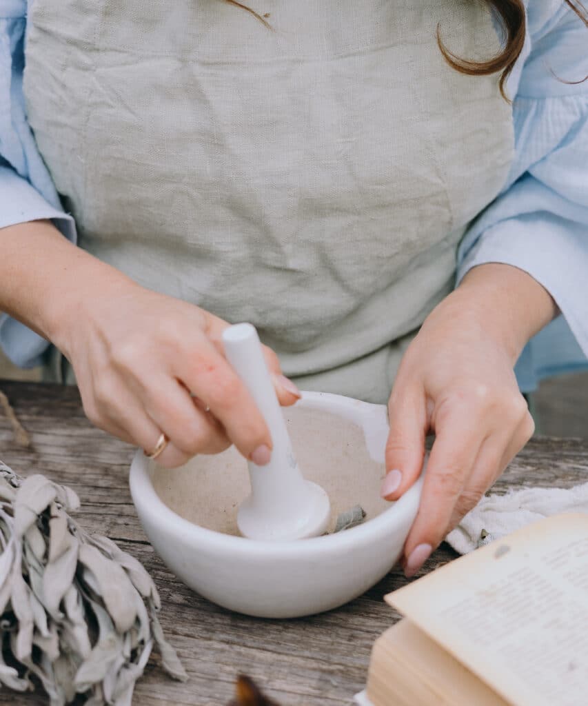 Mains préparant un mélange dans un mortier blanc. Du sage séché et un livre ancien sont posés sur une table en bois rustique.