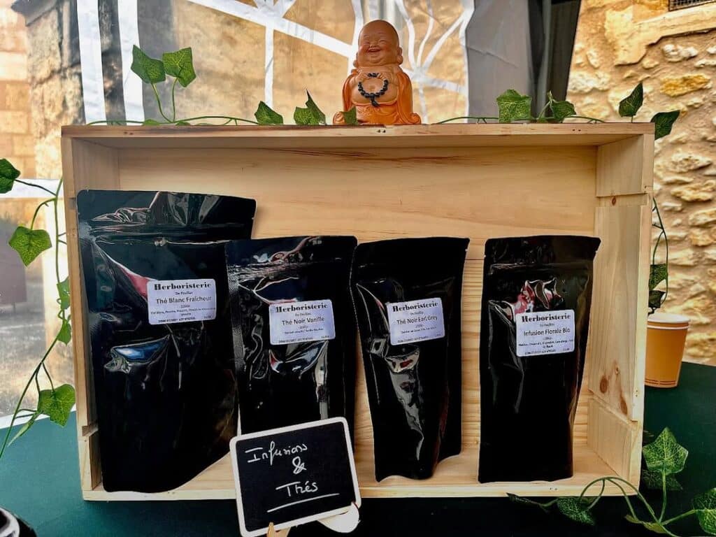 Étalage d'herboristerie : caisse en bois, Bouddha souriant, lierre. Quatre sachets de thés et infusions, dont Thé Blanc Fraîcheur, Thé Noir Earl Grey. Panneau : 'Infusions & Thés'.