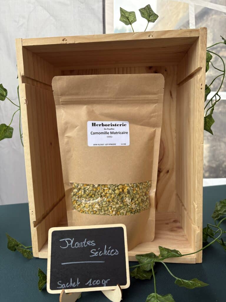 Sachet kraft de 100gr de Camomille Matricaire séchée de l'Herboristerie De Pauillac, prix 12.90€, présenté dans une caisse en bois.